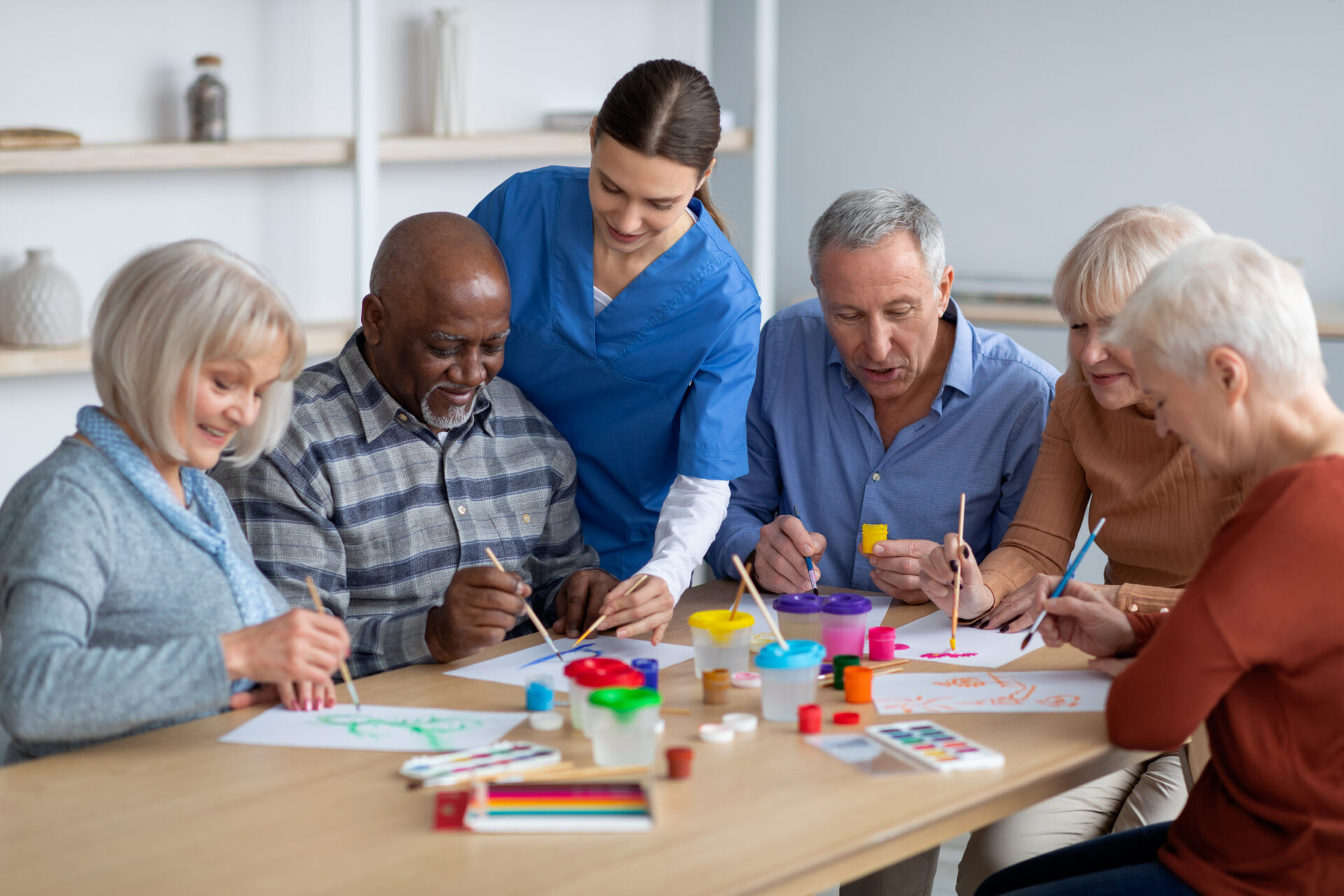 Cheerful creative multiracial senior men and women sitting around table and drawing at nursing home, doing arts and crafts together, young lady nurse assisting group of elderly people, copy space