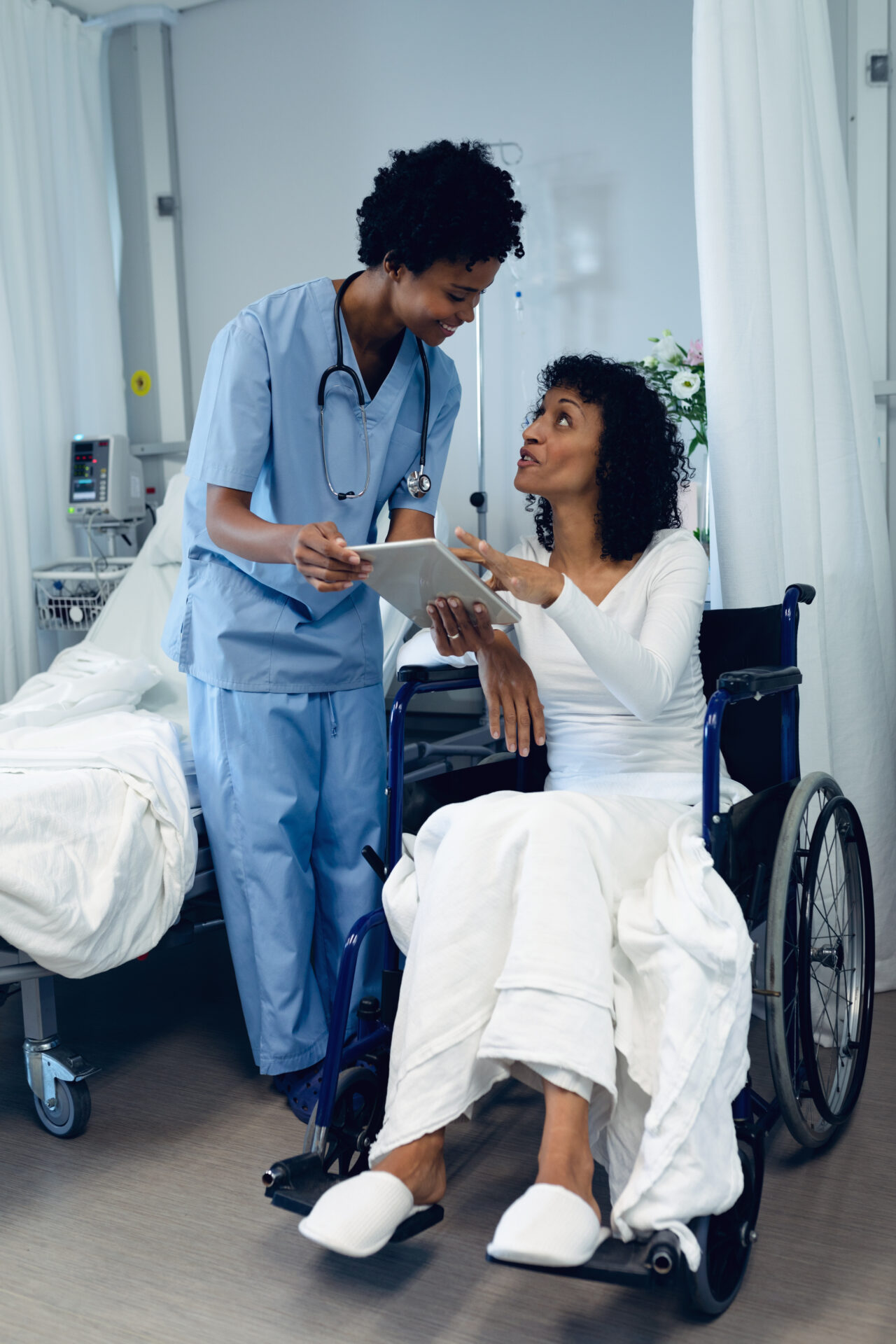 Side view of African-American female doctor helping disabled female patient to use digital tablet in the ward at hospital