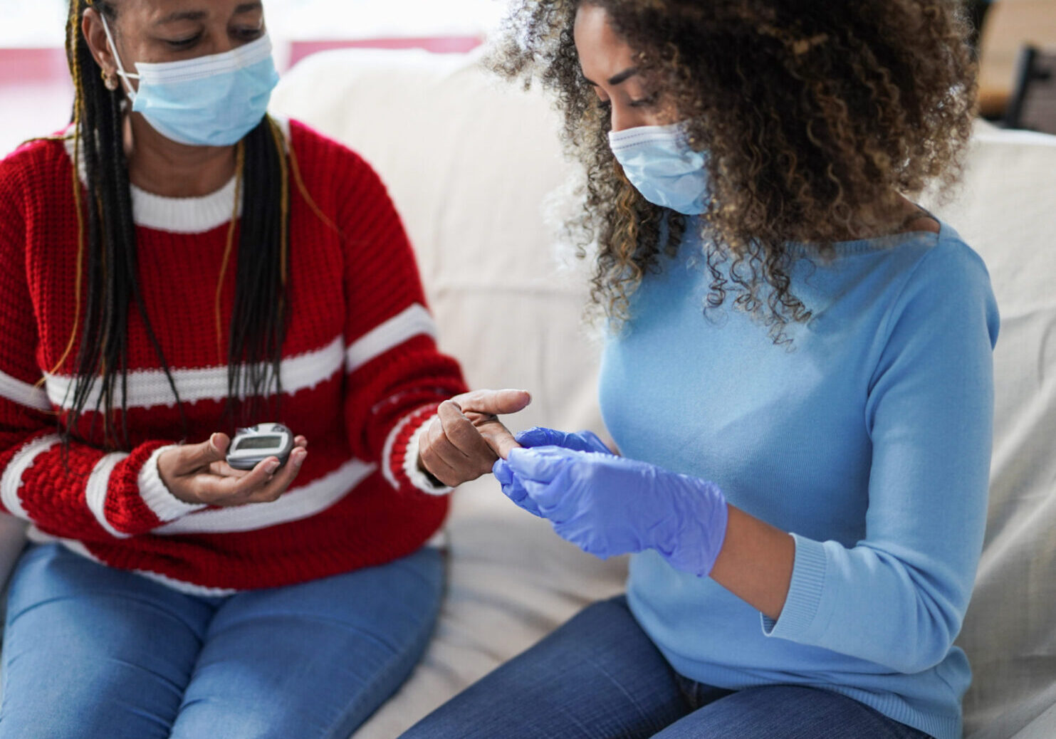 Nurse woman takes blood from a senior african patient at home for the diabetes exam while wearing surgical face mask for coronavirus outbreak